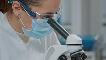A researcher wearing protective safety goggles, a face mask, and a lab coat looks through a microscope lens in a laboratory setting.